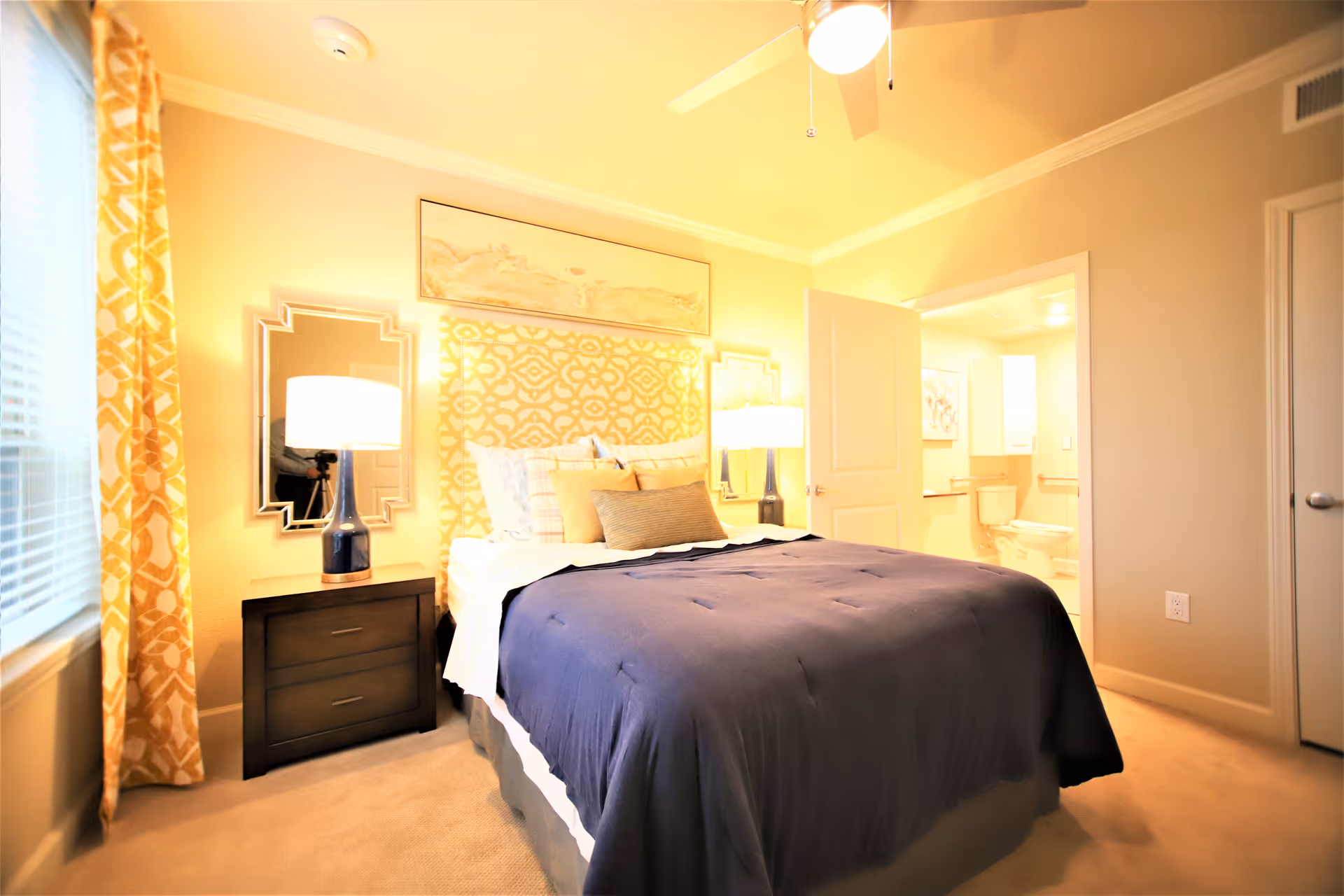 Bright furnished bedroom with a bed covered by a dark duvet, nightstands and lamps, patterned curtains, and an open door leading to a bathroom.