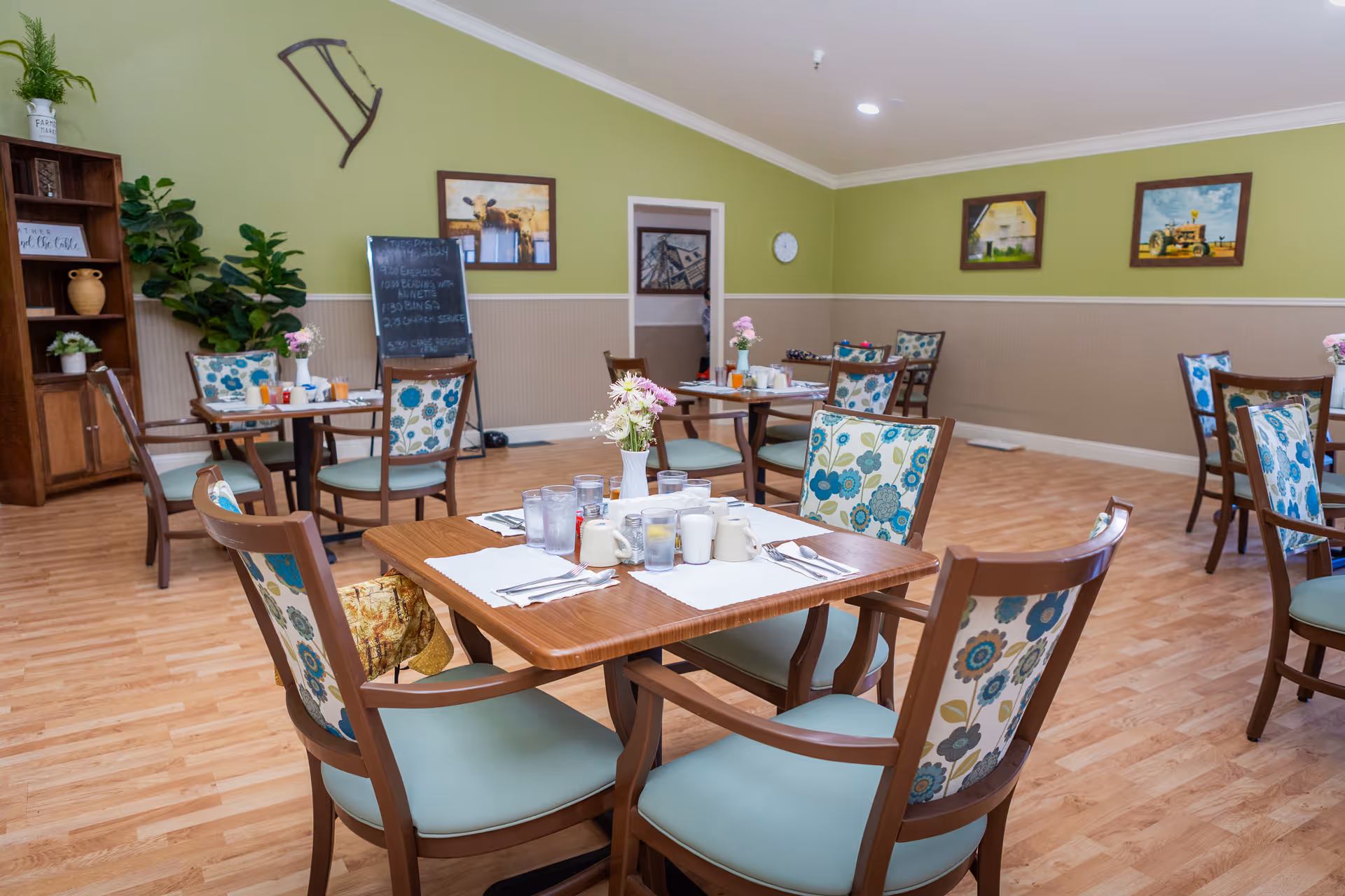 Bright dining room with wooden tables and floral-upholstered chairs set for meals in a senior living facility.