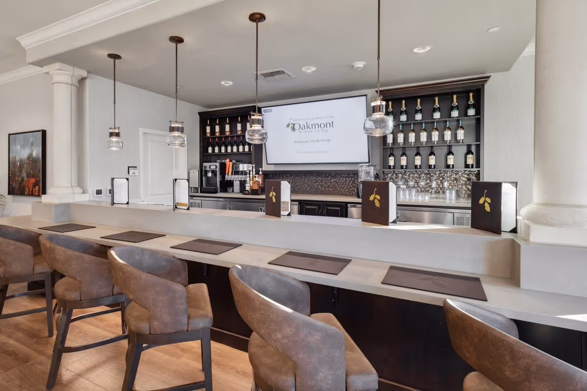 Interior view of a modern bar area with a row of brown cushioned bar stools along a light-colored counter. Behind the counter are shelves stocked with bottles of wine and liquor, a coffee machine, and a large screen displaying the Oakmont Senior Living logo and slogan. The space is well-lit with hanging pendant lights and has a clean, contemporary design.