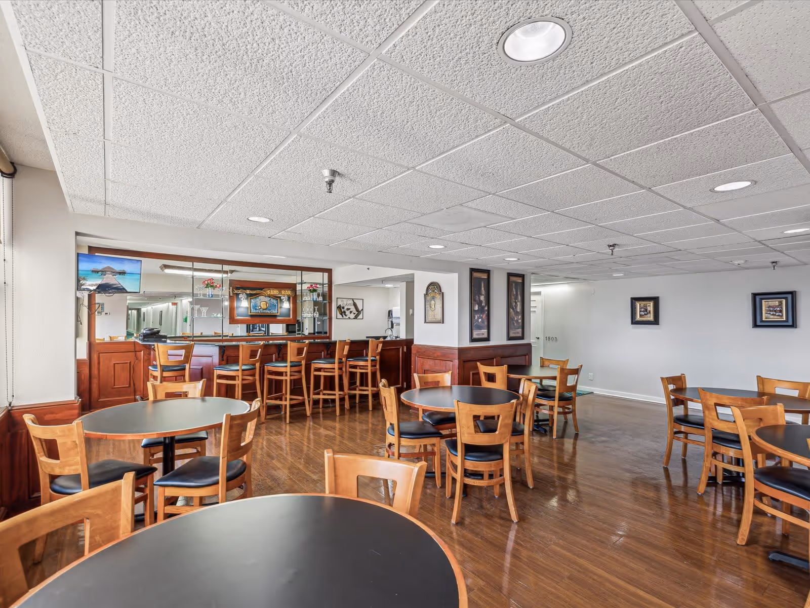 Interior view of a dining area with multiple round tables and wooden chairs with black seats. There is a bar counter with high wooden stools and a mirror behind it. The floor is wooden, and the ceiling has white acoustic tiles with recessed lighting. The walls are decorated with framed pictures.