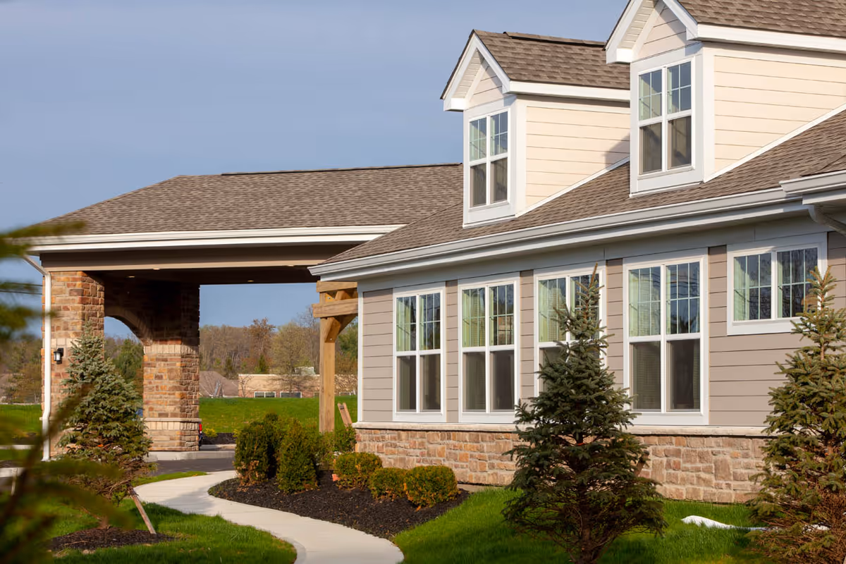 Exterior view of a senior living facility building with beige siding, multiple windows, and a stone base. There is a covered entrance supported by stone pillars, a curved sidewalk, and landscaped greenery including small pine trees and bushes.