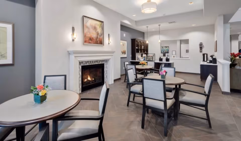 Bright dining area with round tables and upholstered chairs facing a lit fireplace and a service counter.