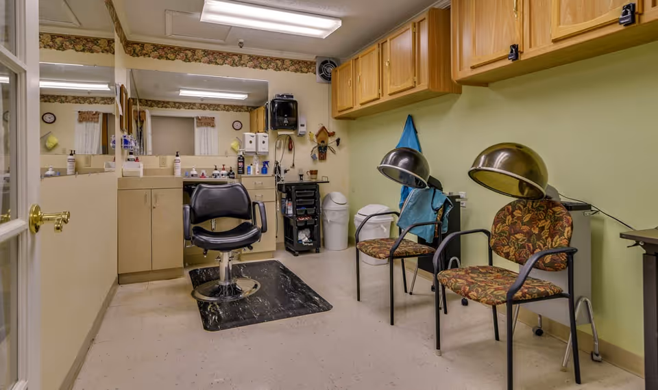 Interior view of a hair salon area in a nursing center with a black salon chair on a black mat in front of a counter with a large mirror. To the right, there are two floral-patterned chairs with vintage hair dryers attached. The walls are light green with wooden cabinets mounted above the chairs.