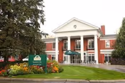 Front exterior view of Westminster Village North, a large brick building with white columns and a triangular pediment above the entrance. There is a green sign with the facility name surrounded by colorful flowers and greenery in the foreground.