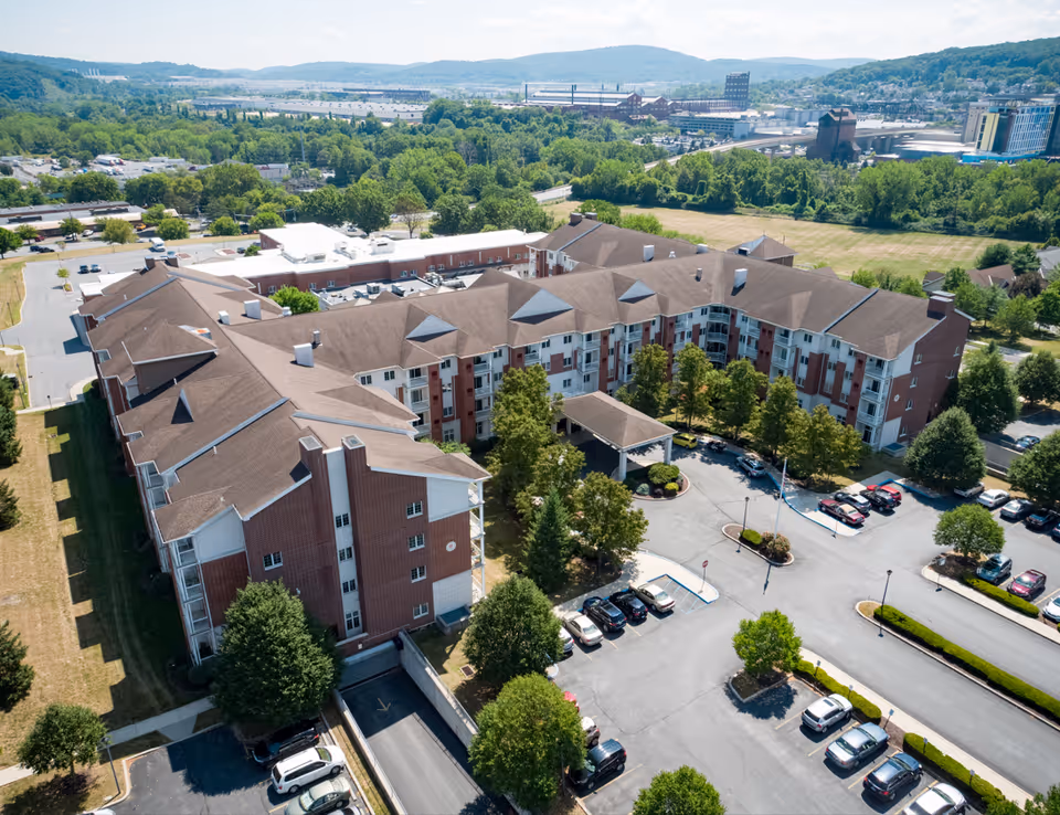 Aerial view of Moravian Village of Bethlehem, showing a large multi-story senior living facility with brown roofs and red brick walls surrounded by trees and parking lots. The background includes a cityscape with hills and various buildings under a partly cloudy sky.