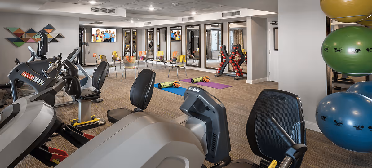 A well-equipped fitness room with exercise bikes, yoga mats, small basketballs, chairs arranged in a semi-circle, a rack of dumbbells, and large exercise balls stacked on the right side. The room has large mirrors on one wall and a TV mounted above the chairs.