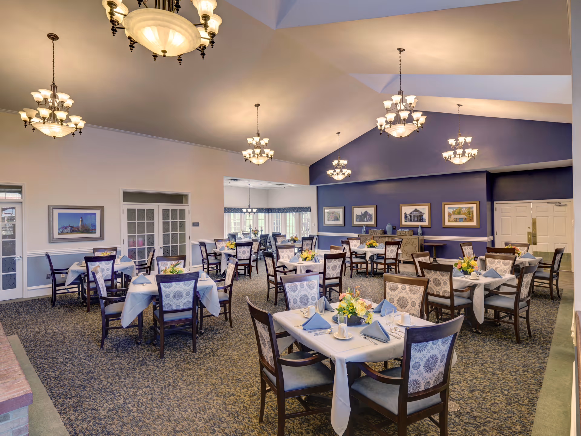 A spacious dining room with multiple tables covered in white tablecloths, each set with blue napkins, cups, and floral centerpieces. The room features high ceilings with elegant chandeliers, patterned carpet, and framed artwork on the walls. Large windows allow natural light to brighten the space.
