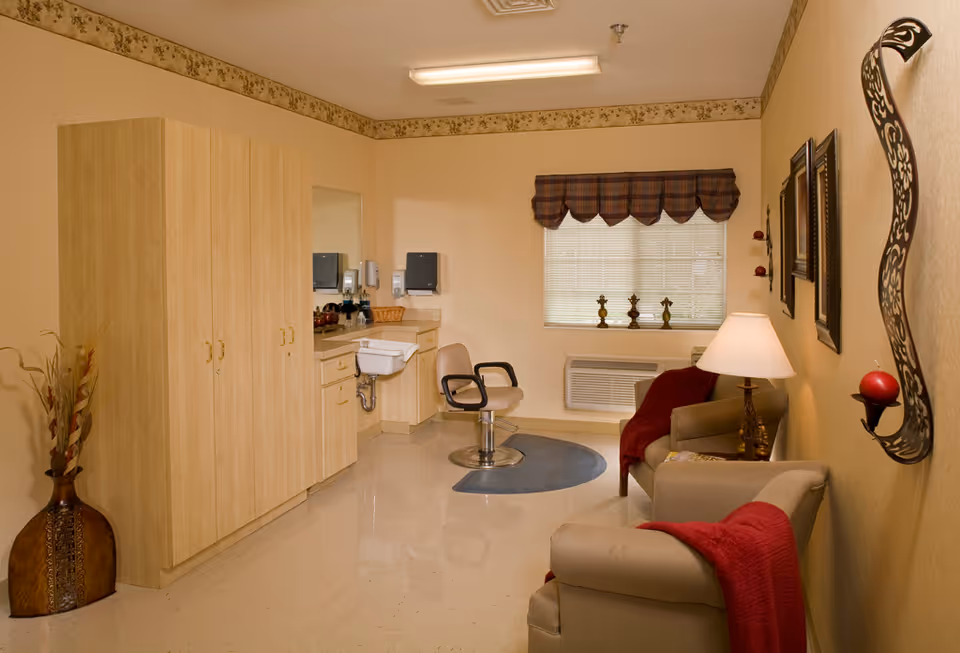 A cozy room with beige walls and floor, featuring a large wooden cabinet, a small sink with countertop and mirror, a swivel chair on a circular rug, two armchairs with red throws, a table lamp, decorative wall sconces, and a window with a plaid valance.