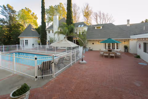 Outdoor patio area of a senior living facility with a fenced swimming pool, patio furniture including a table with an umbrella, and a large building with multiple windows and a chimney surrounded by trees.