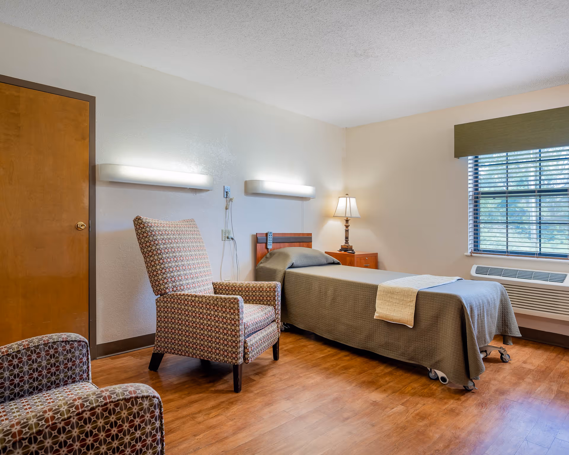 A simple, clean bedroom in a care facility featuring a single bed with a green bedspread and a beige throw, a wooden nightstand with a lamp, two patterned armchairs, a window with blinds and a green valance, and a wooden door. The room has wood flooring and white walls with two wall-mounted lights above the bed.