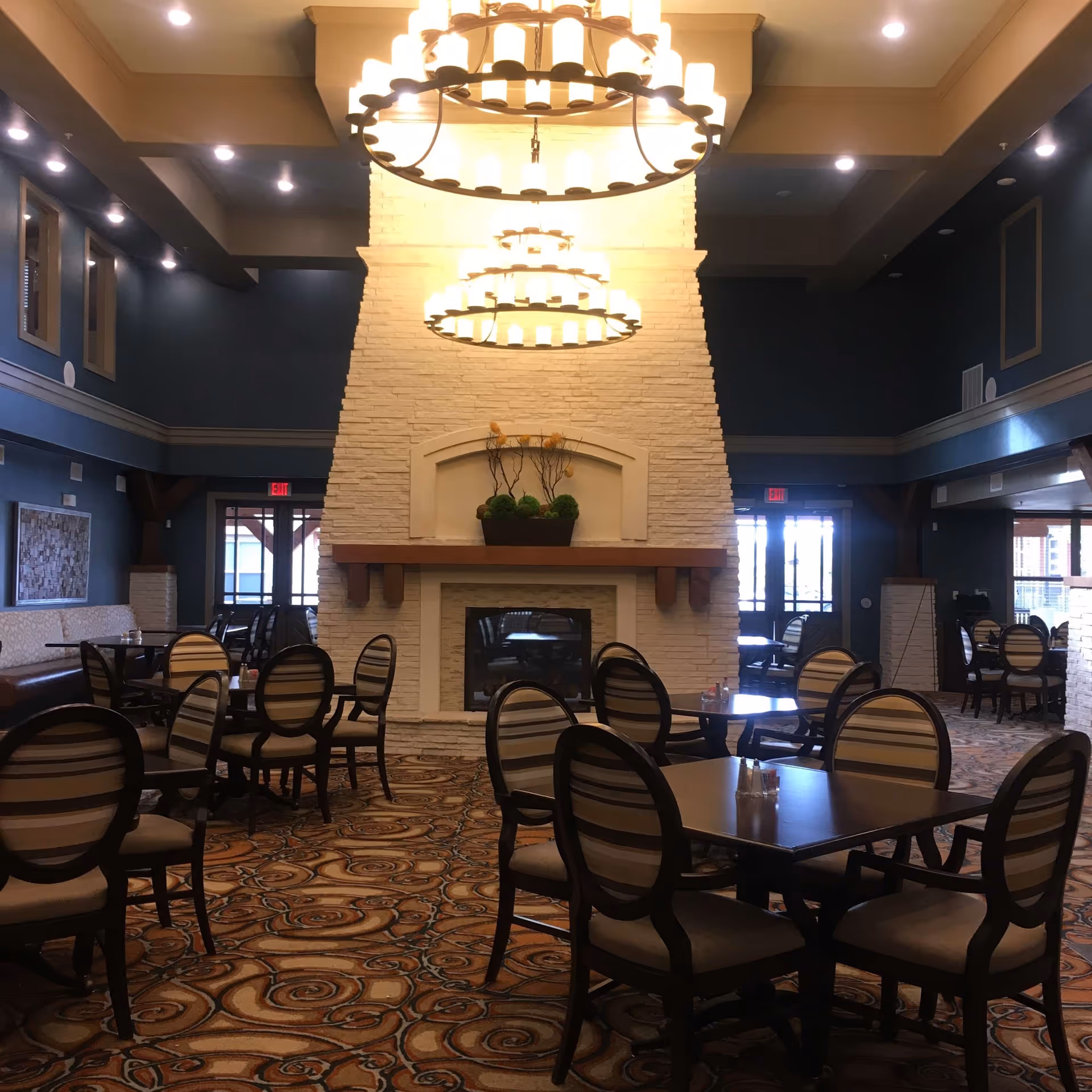 A spacious dining area with multiple tables and chairs arranged on a patterned carpet. The room features a large white stone fireplace in the center with decorative plants on the mantel. Two large circular chandeliers hang from the ceiling, illuminating the space. The walls are painted dark blue with several windows and exit doors visible in the background.