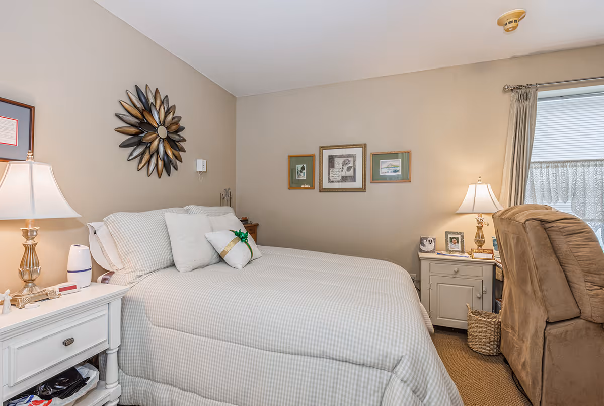 A cozy bedroom with a neatly made bed covered in light-colored checkered bedding. There are two bedside tables, each with a lamp, and framed pictures hanging on the beige walls. A brown recliner chair is positioned near a window with curtains and blinds.