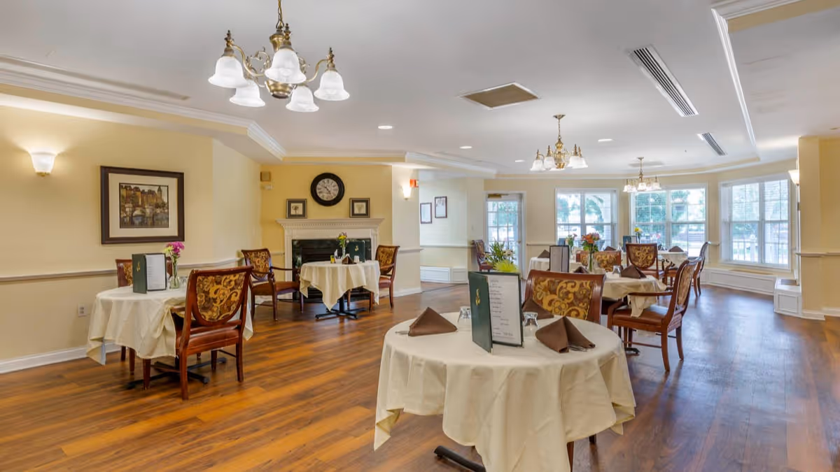 A bright and spacious dining room with several round tables covered with white tablecloths and set with menus and folded brown napkins. The room features wooden flooring, large windows allowing natural light, elegant chandeliers, and a fireplace with a clock and framed pictures above it. The walls are painted a soft yellow, and the chairs have ornate upholstery.