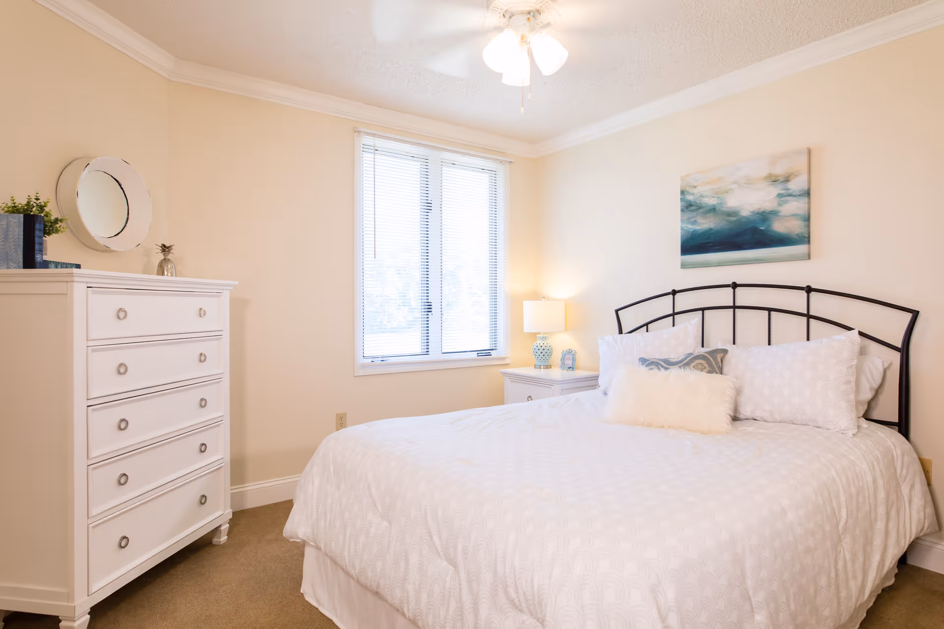 Bright bedroom with a bed, white dresser, nightstand with a lamp, and a window.