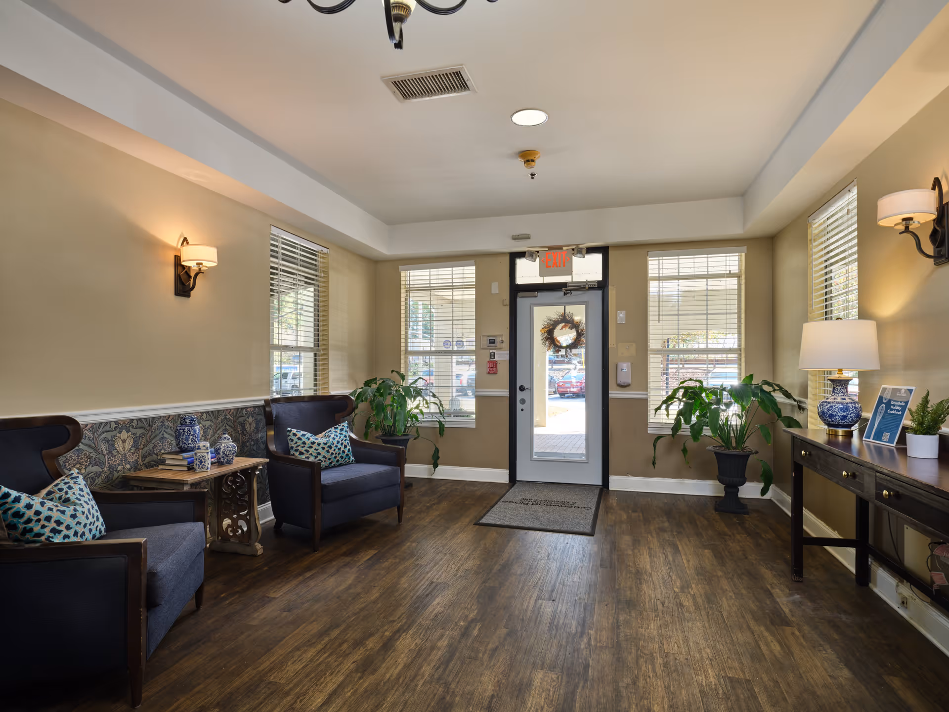A cozy assisted living facility lobby with two dark armchairs featuring patterned cushions, a small wooden side table with decorative items, potted plants, a wooden console table with a blue and white lamp, and a glass door with windows on either side letting in natural light.