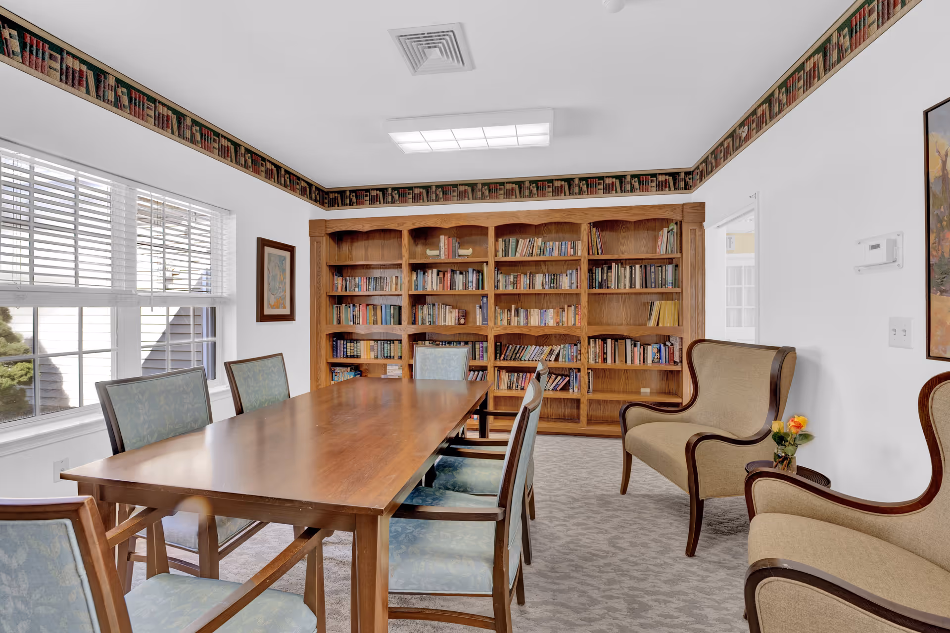A bright room with a large wooden table surrounded by six chairs with light blue upholstery. Against the back wall is a large wooden bookshelf filled with books. Two beige armchairs with wooden frames are positioned to the right, with a small table holding a vase of flowers between them. The room has white walls, a window with blinds on the left, and a decorative wallpaper border featuring books near the ceiling.