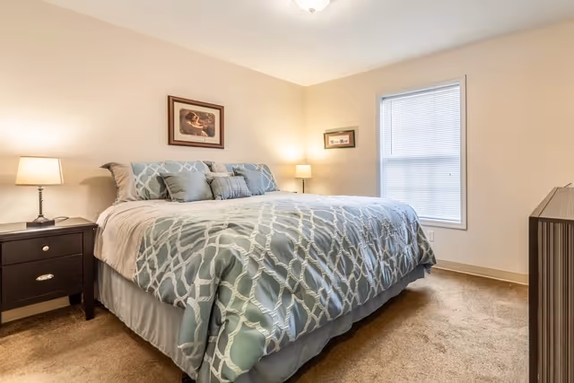 A bedroom with a large bed covered in a blue and white patterned comforter and multiple pillows. There are two nightstands on either side of the bed, each with a lamp. A window with blinds is on the right wall, and two framed pictures hang on the walls. The room has beige carpet and light-colored walls.