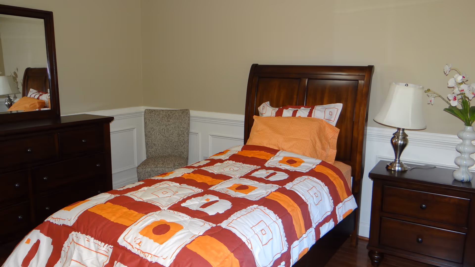 A bedroom with a single wooden bed featuring a colorful orange, red, and white patterned comforter and pillows. To the right of the bed is a wooden nightstand with a silver lamp and a white vase with flowers. To the left is a wooden dresser with a large mirror and a patterned upholstered chair in the corner. The walls are beige with white wainscoting.