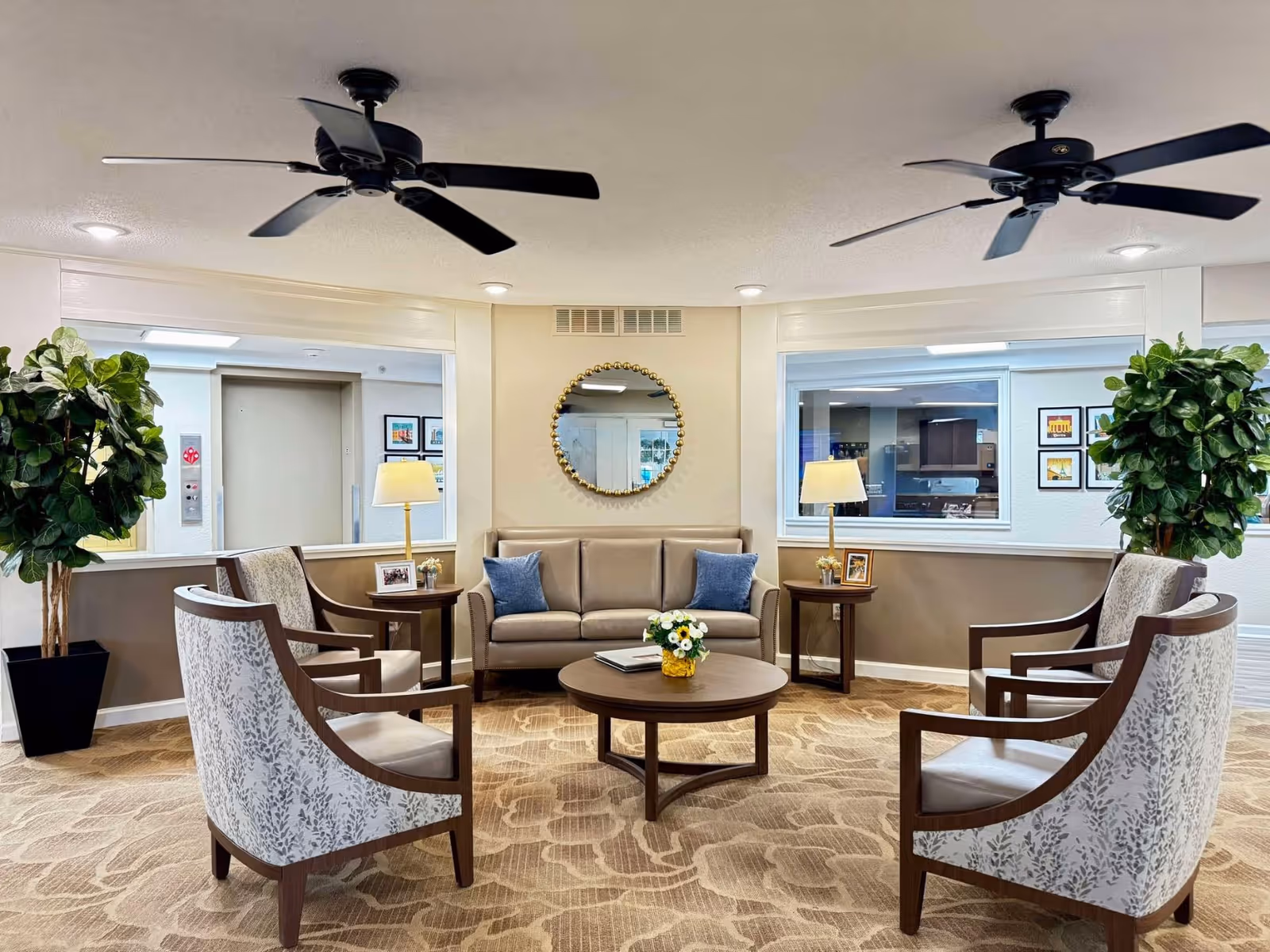 A cozy seating area in a senior living facility with a beige sofa adorned with two blue pillows, four patterned armchairs arranged around a round wooden coffee table with a flower arrangement, two side tables with lamps and framed photos, two large potted plants, a round decorative mirror on the wall, and two ceiling fans above.