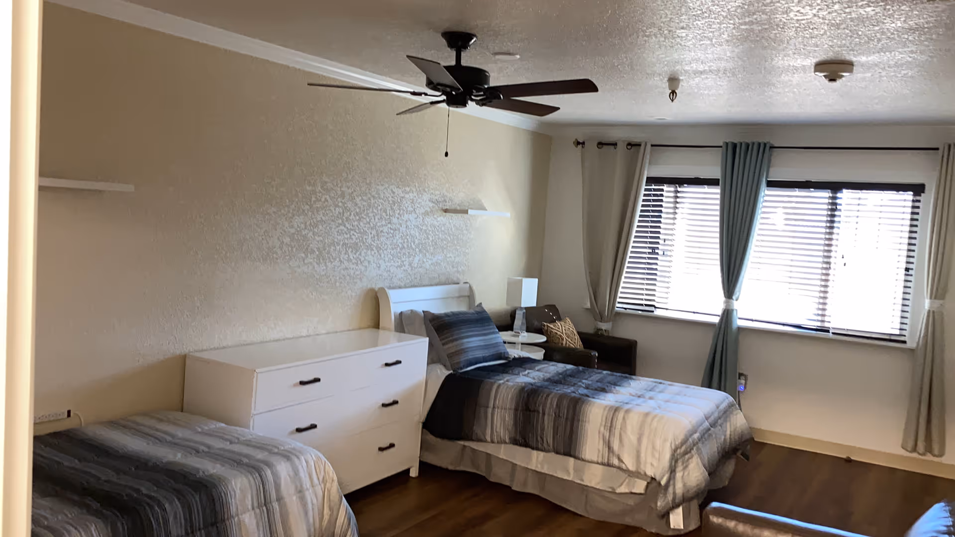 A bedroom with two single beds, each with striped bedding in shades of gray and blue. Between the beds is a white dresser with three drawers and a small round white side table with a lamp. There is a brown armchair near a window with blinds and curtains. A ceiling fan with dark blades is mounted on the ceiling. The room has wooden flooring and beige walls.