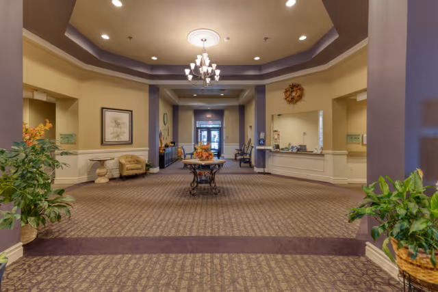 Spacious senior living facility lobby with patterned carpet, a central table decorated with flowers, beige walls with white wainscoting, a chandelier hanging from a recessed ceiling, potted plants on either side, seating areas with chairs and small tables, and a reception desk on the right side.
