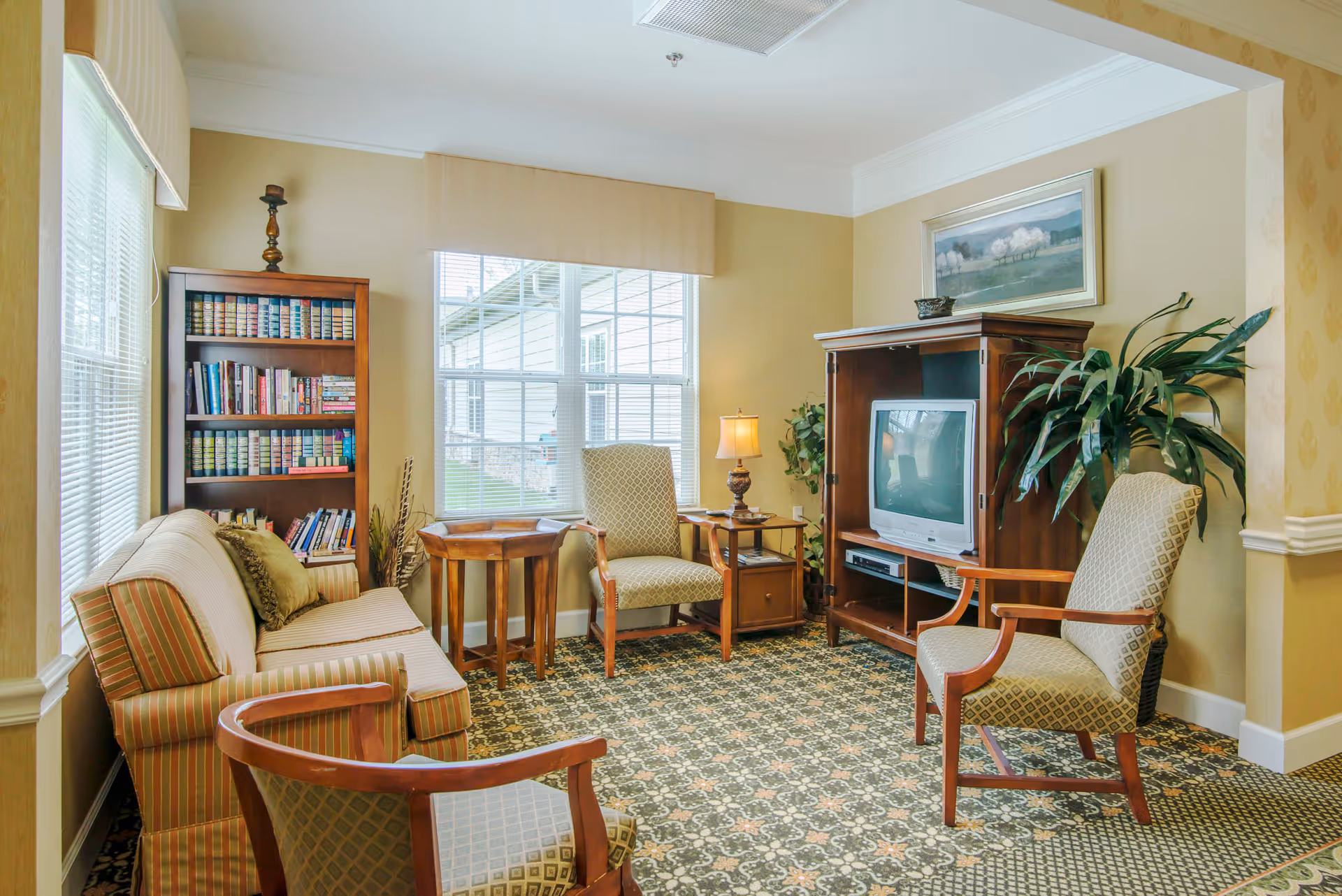 A cozy living room with patterned carpet, a striped sofa, two upholstered armchairs, a wooden bookshelf filled with books, a wooden TV cabinet with an older-style television, a side table with a lamp, a large window with blinds, and a potted plant in the corner.