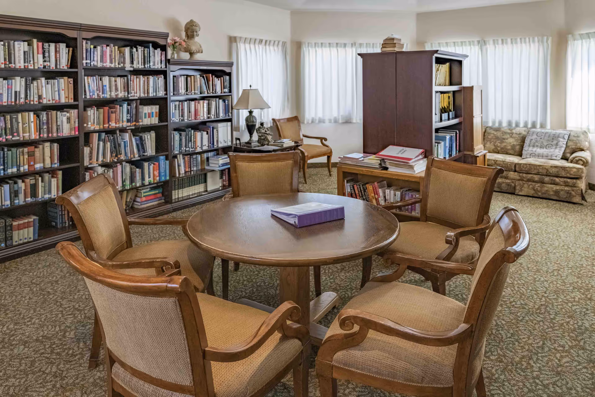 A cozy library room with a round wooden table surrounded by five cushioned wooden chairs. Bookshelves filled with books line the walls, and there is a small lamp and decorative bust on a side table. A patterned sofa sits near windows with white curtains, and the room has a carpeted floor.