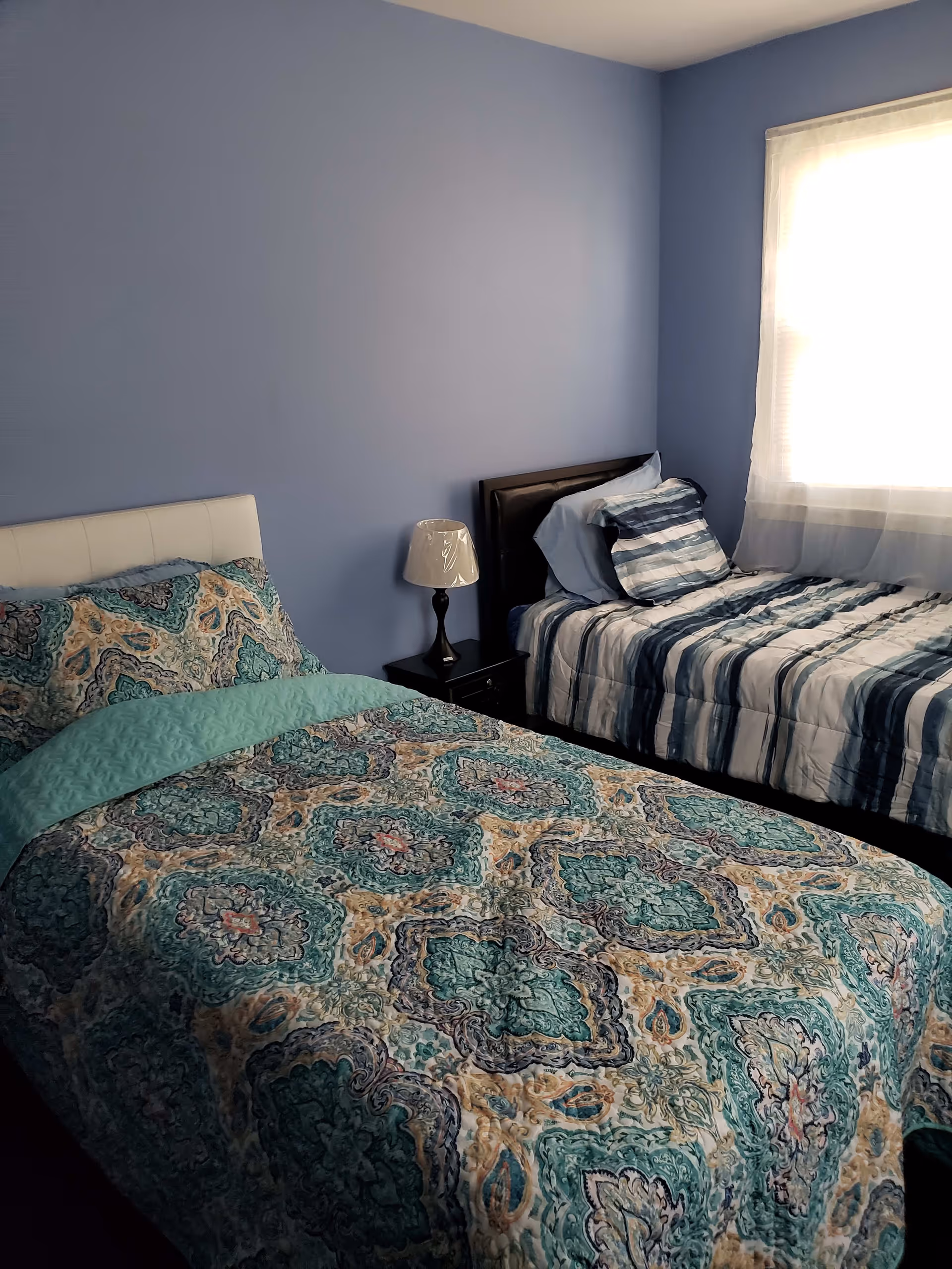 A bedroom with two twin beds, one with a patterned teal, beige, and blue bedspread and the other with a striped blue and white bedspread. There is a small black nightstand between the beds with a lamp on it. The walls are painted light blue and a window with sheer white curtains lets in natural light.