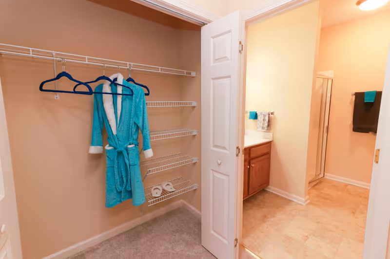Open closet with wire shelving and a teal bathrobe hanging on a hanger, adjacent to a bathroom with a vanity, towels, and a glass shower enclosure.