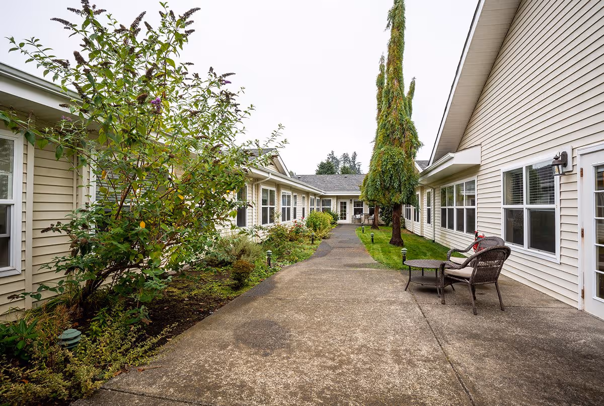Outdoor walkway between two beige buildings with windows, surrounded by green shrubs and trees, with a small seating area consisting of two wicker chairs and a table on the right side.