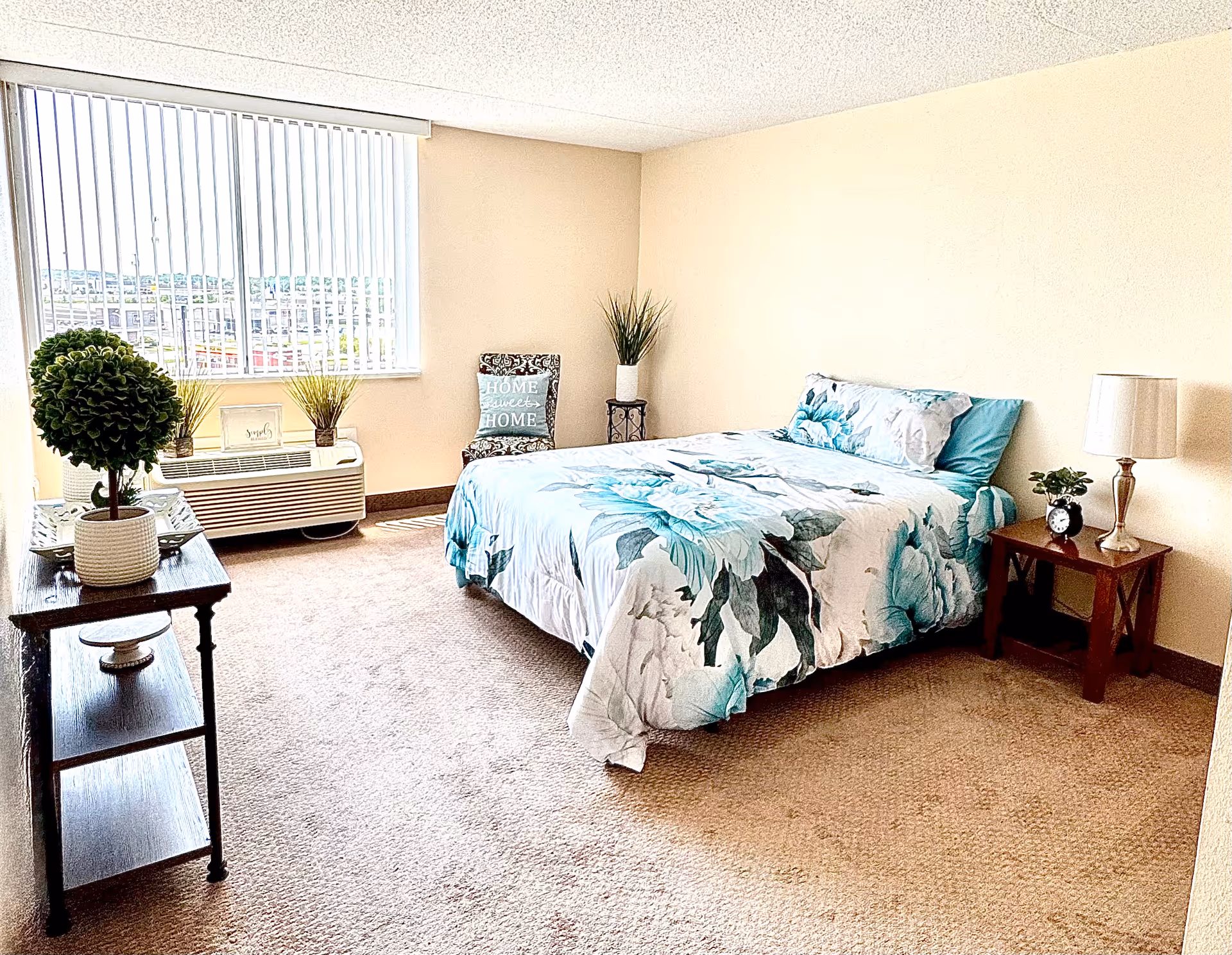 A bright bedroom with a large window covered by vertical blinds. The room features a bed with floral bedding in shades of teal and white, a wooden nightstand with a lamp and small plant, a decorative chair with a cushion that says 'HOME sweet HOME', and a wooden table with a potted plant. The walls are light beige and the floor is carpeted.