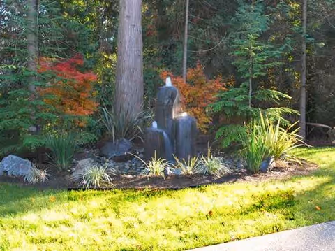 Outdoor garden with a small waterfall statue, surrounding rocks, and lush shrubs on a sunny lawn.