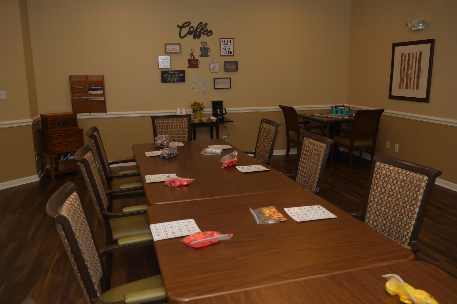 Communal dining/activity room with a long wooden table set with bingo cards and snack bags, surrounded by chairs and wall decor reading 'Coffee'.