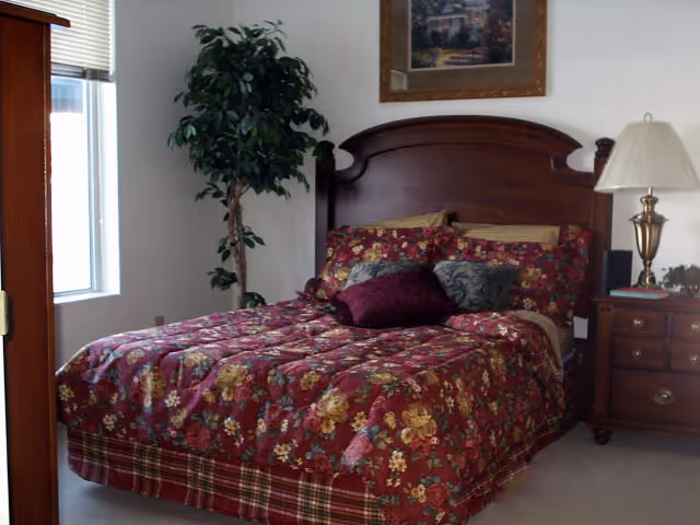 A bedroom with a wooden bed frame and a floral-patterned bedspread in shades of red, yellow, and green. The bed has multiple pillows, including a burgundy one. To the right of the bed is a wooden nightstand with a brass lamp and some decorative items. A framed picture hangs on the wall above the bed, and there is a tall green plant near a window with blinds on the left side of the room.