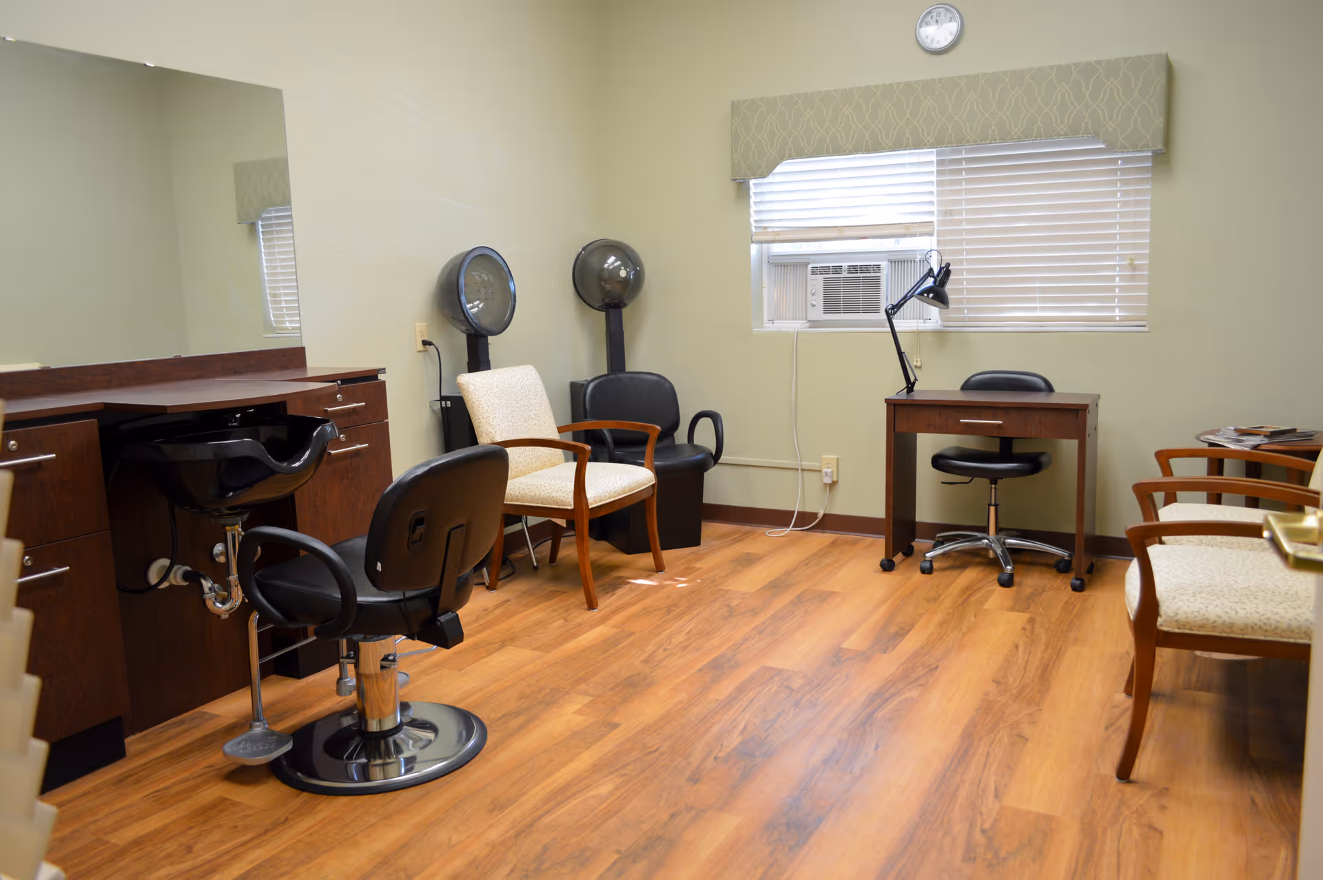 Small salon-style room with styling chairs, hooded hair dryers, a sink and mirror, a desk, and waiting chairs on wood flooring.