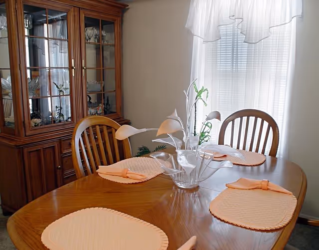 A wooden dining table set with four peach-colored placemats and matching napkins, accompanied by wooden chairs. A decorative glass vase with white calla lily flowers is placed in the center of the table. In the background, there is a wooden china cabinet with glass doors displaying glassware, and a window with sheer white curtains allowing natural light into the room.