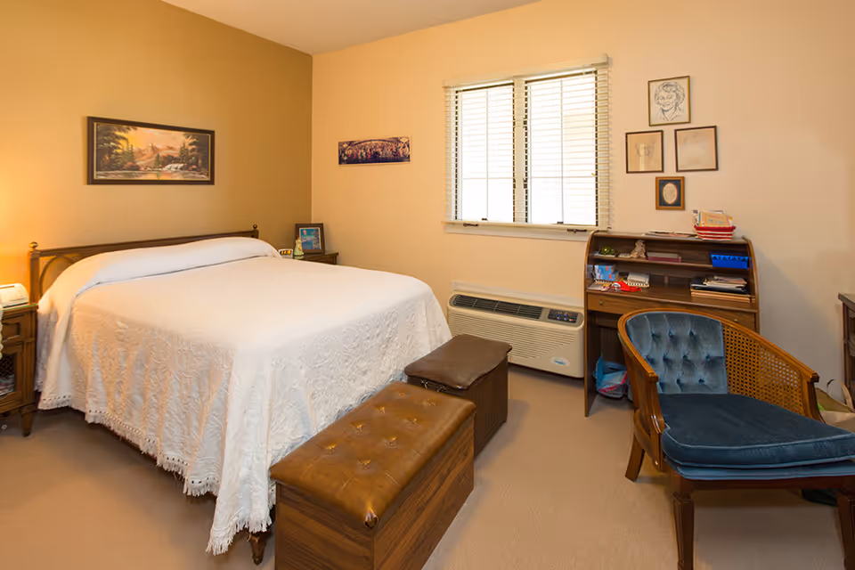A cozy bedroom with a large bed covered in a white textured bedspread. There is a wooden nightstand with a lamp and a phone on the left side of the bed. At the foot of the bed are two brown leather storage benches. On the right side of the room, there is a wooden roll-top desk with various items on it and a blue cushioned wooden chair. The walls are decorated with framed pictures and a window with white blinds is centered on the wall behind the desk.