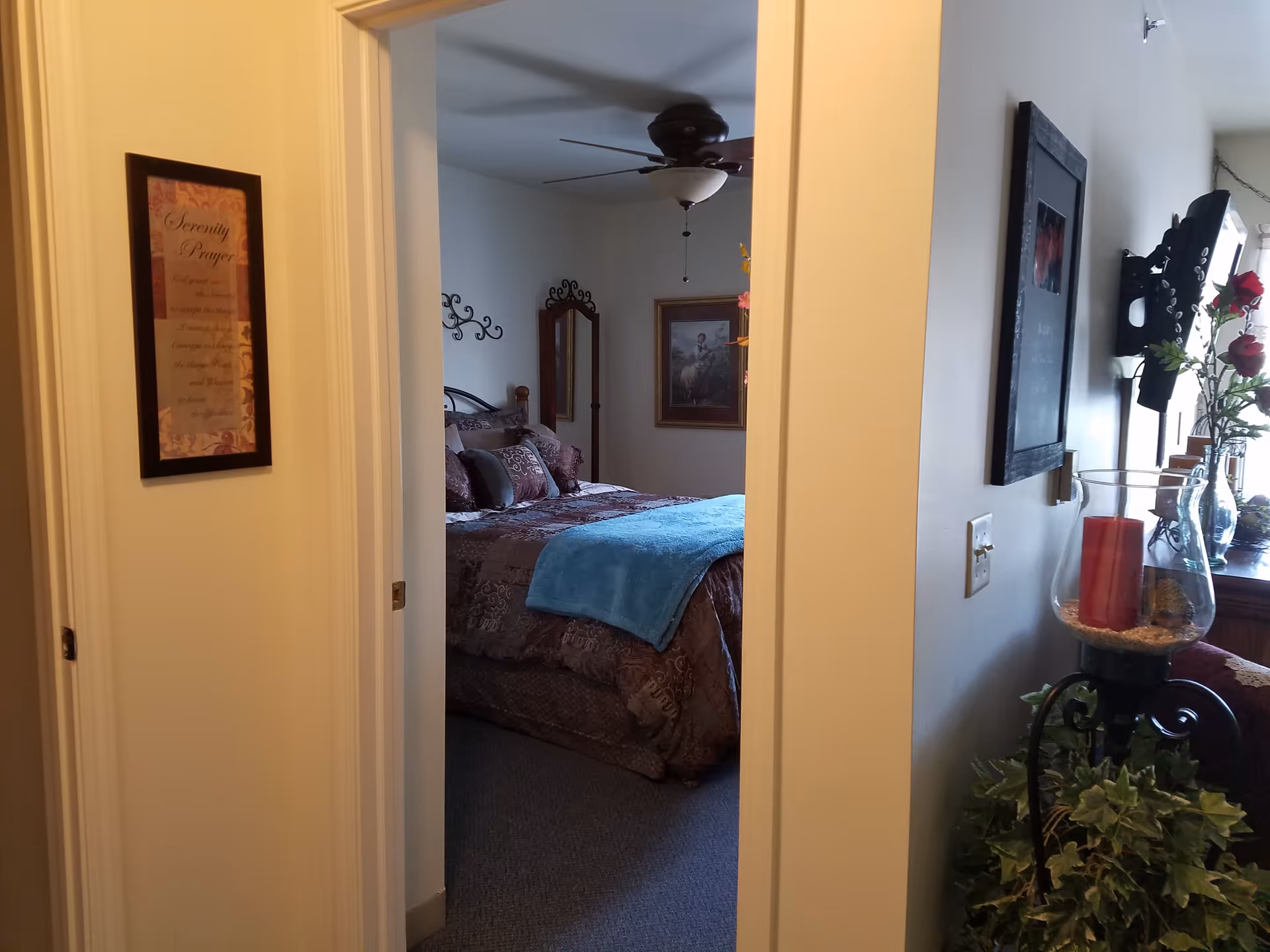 View through a doorway into a bedroom with a bed covered in patterned bedding and a blue throw blanket. The bedroom has a ceiling fan, wall decorations including a framed picture and a mirror. The hallway outside the bedroom has framed wall art and a decorative candle holder with a red candle and greenery.