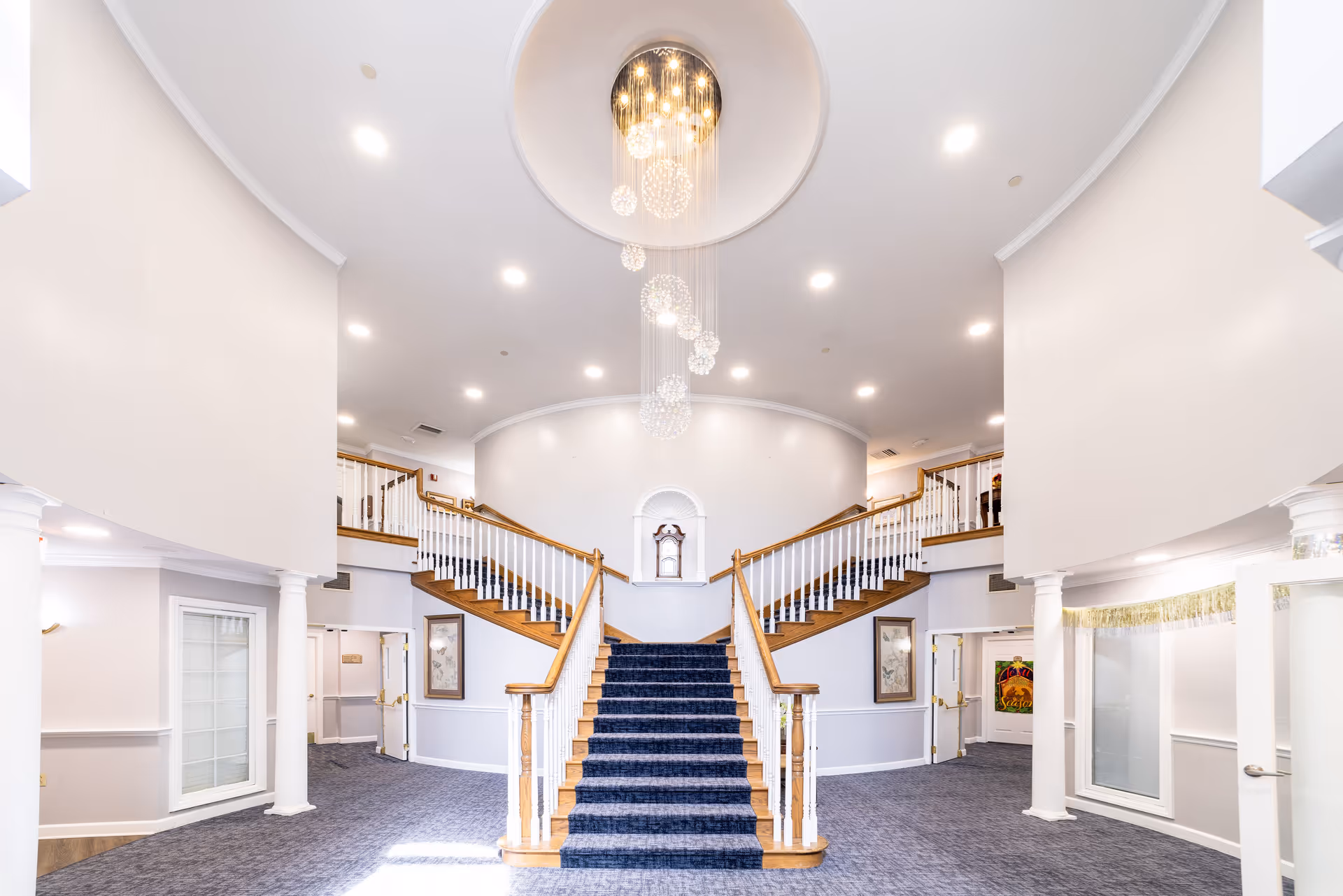 Bright and spacious interior of a senior living facility featuring a grand staircase with blue carpet leading to an upper level. The staircase splits into two directions at the top. The walls are painted white with wooden handrails and white balusters. A modern chandelier with multiple hanging light spheres is centered above the staircase. The area is well-lit with recessed ceiling lights and has carpeted floors. There are framed pictures on the walls and white columns supporting the upper level.