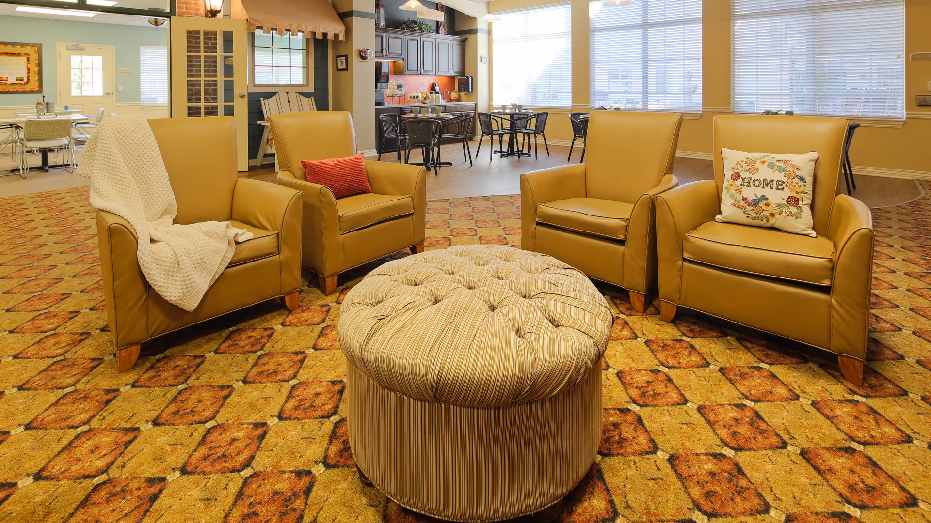 A cozy common area with four tan leather armchairs arranged around a large round tufted ottoman on a patterned carpet. Two chairs have decorative pillows, one with the word 'HOME' and another with a red pillow. In the background, there are tables and chairs near large windows with blinds, and a kitchen area with cabinets and appliances.