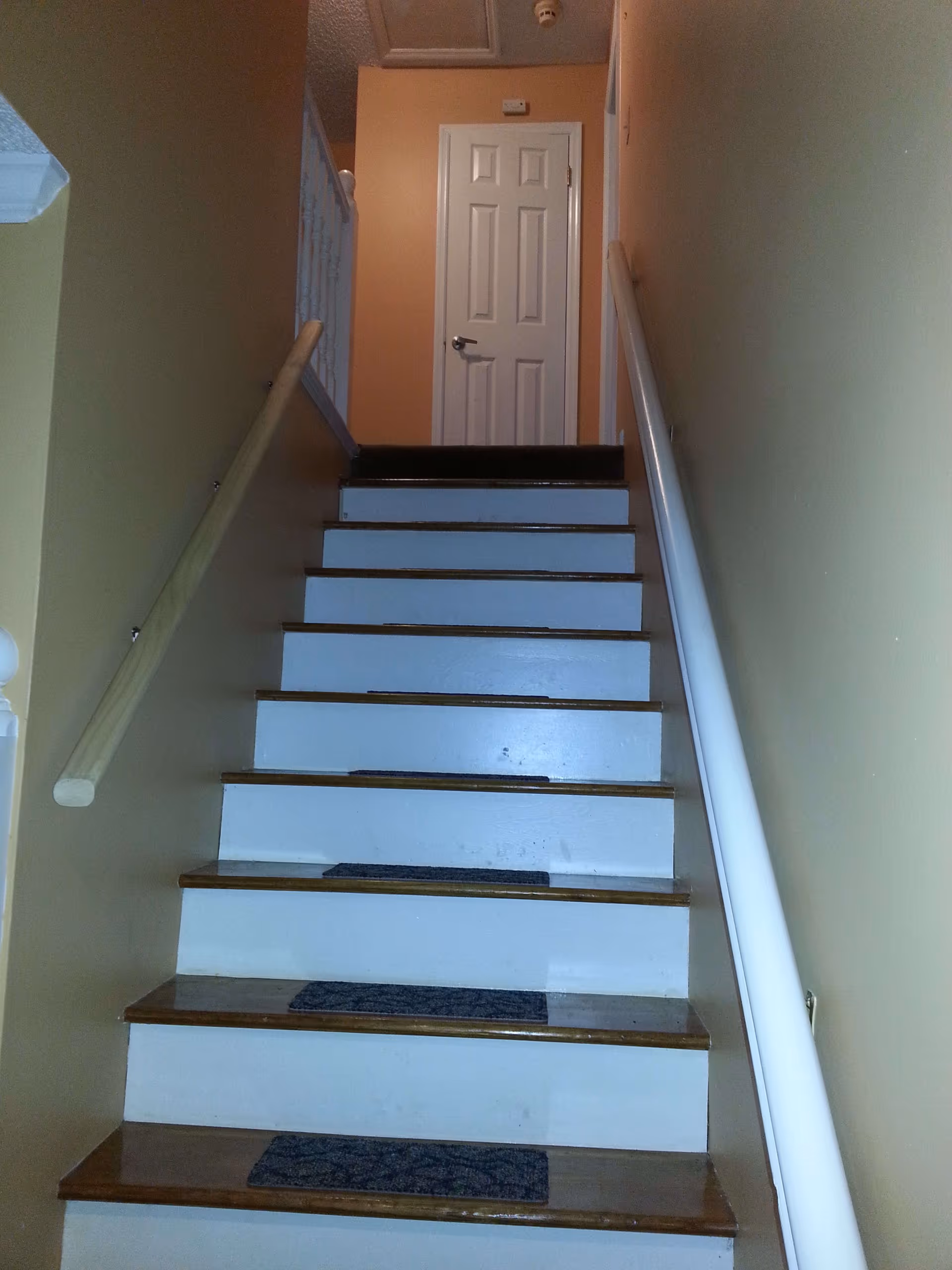 A staircase with wooden steps and white risers leading up to a closed white door. There are handrails on both sides of the staircase and beige walls surrounding the area.
