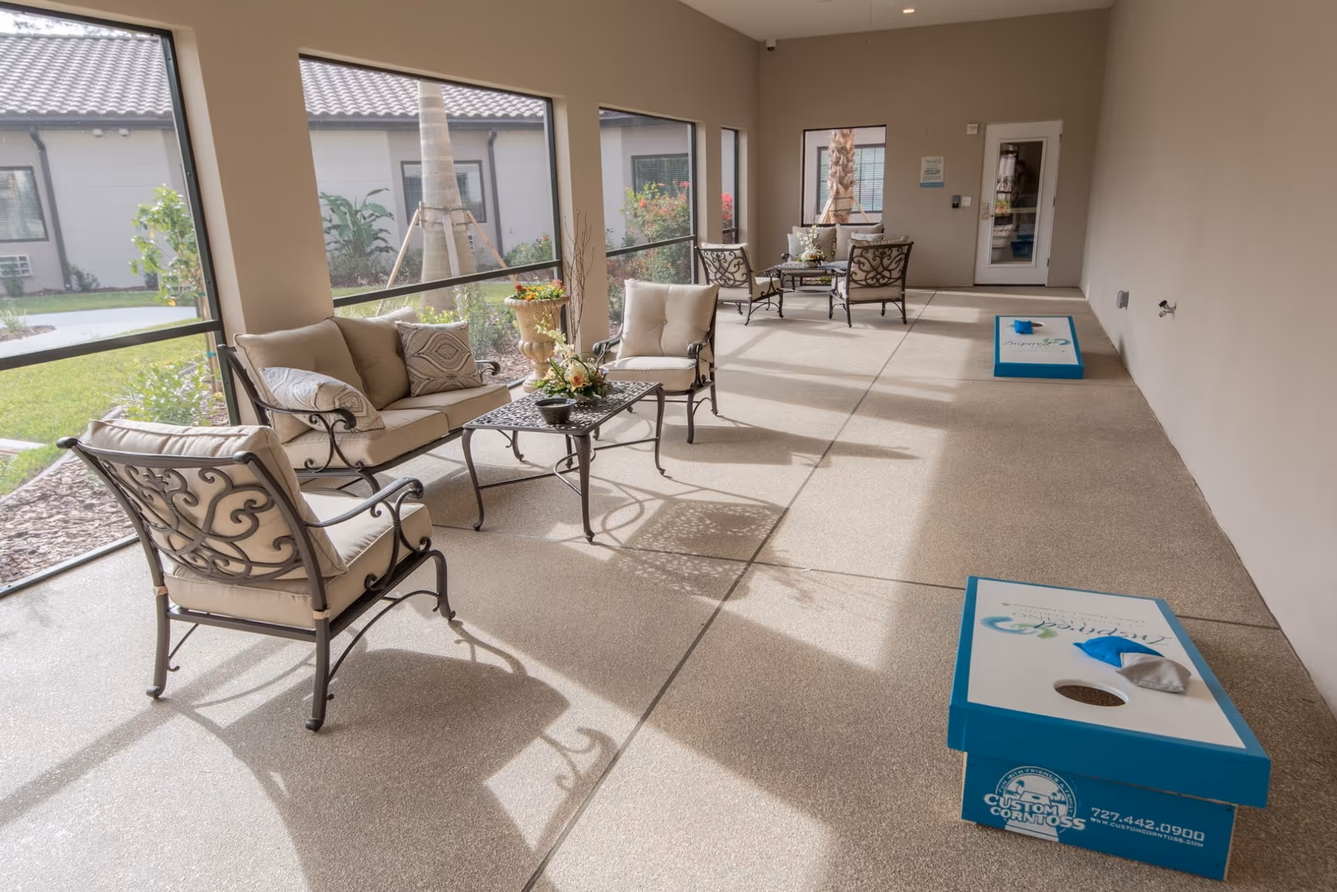 Sunlit enclosed patio with cushioned metal seating, small tables, and two cornhole boards.