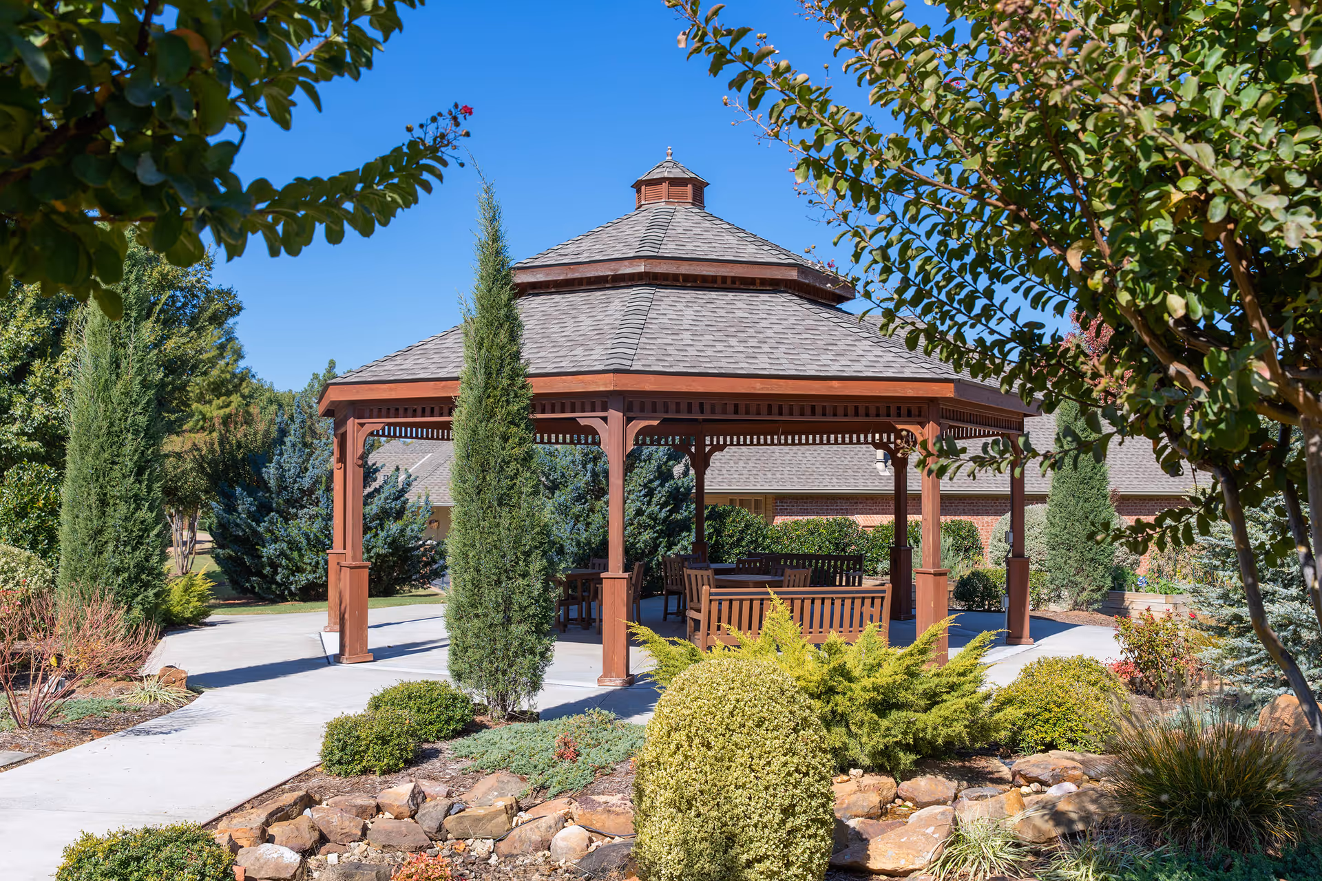 A wooden gazebo with a shingled roof surrounded by landscaped greenery including bushes, small trees, and rocks under a clear blue sky. There are benches and tables inside the gazebo, and a concrete pathway leads to it.
