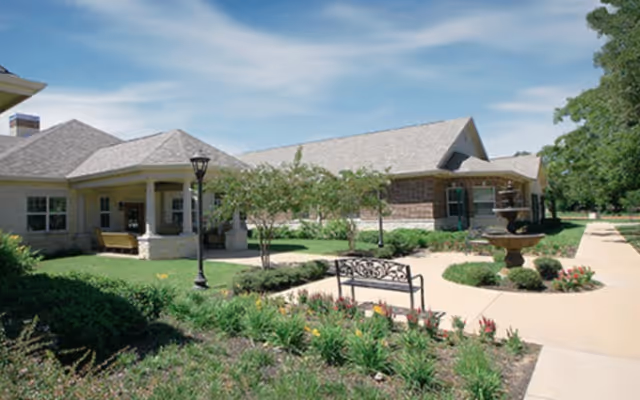 Outdoor view of a senior living facility with a paved walkway, landscaped garden beds with flowers and shrubs, a bench, a lamp post, and a water fountain. The building has a light-colored exterior with a covered porch area and multiple windows under a partly cloudy sky.