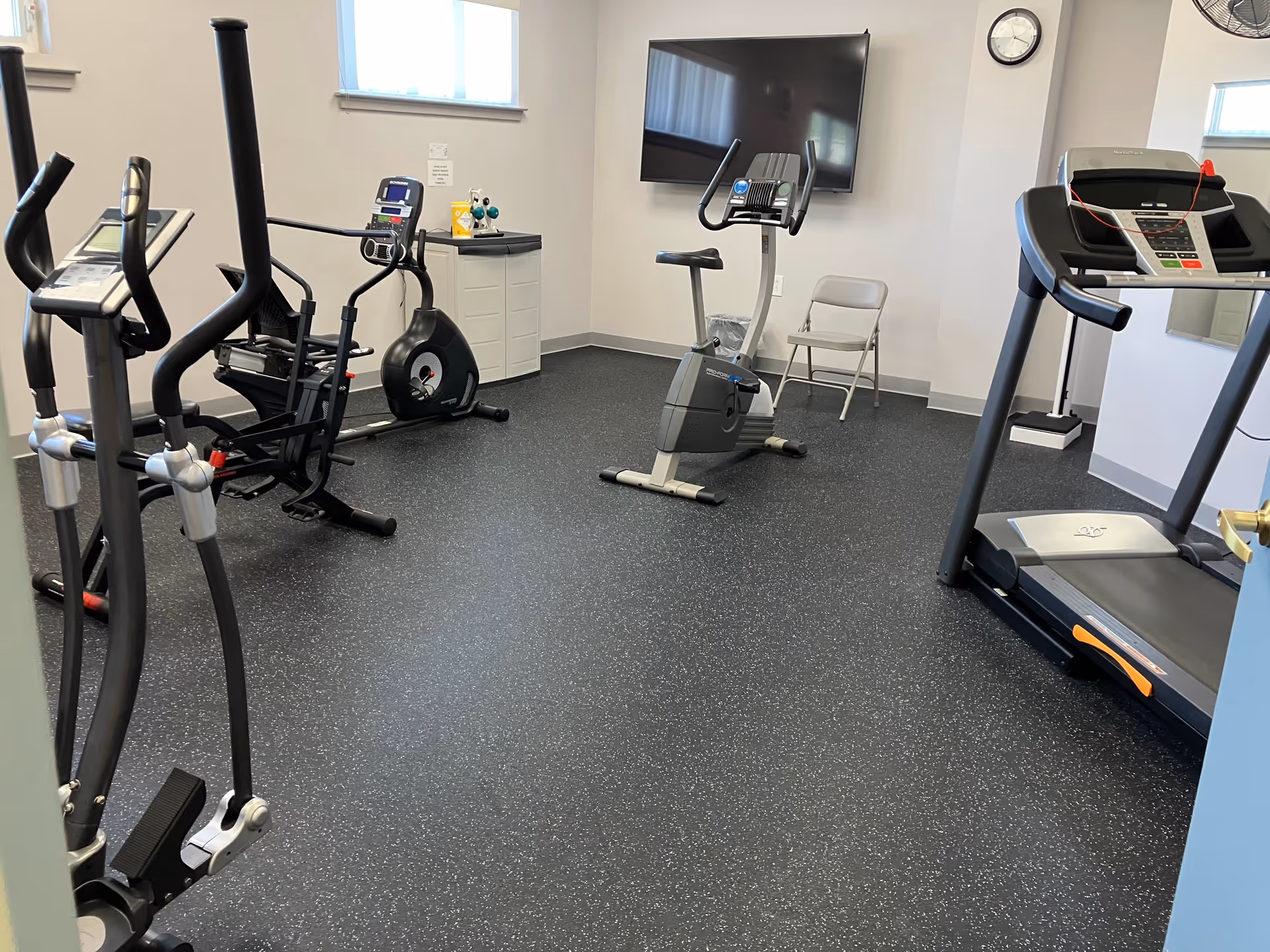 A small fitness room with exercise equipment including an elliptical machine, a stationary bike, a treadmill, and a rowing machine. There is a folding chair, a wall-mounted TV, a clock, and a small cabinet with cleaning supplies. The room has two windows and a speckled black rubber floor.
