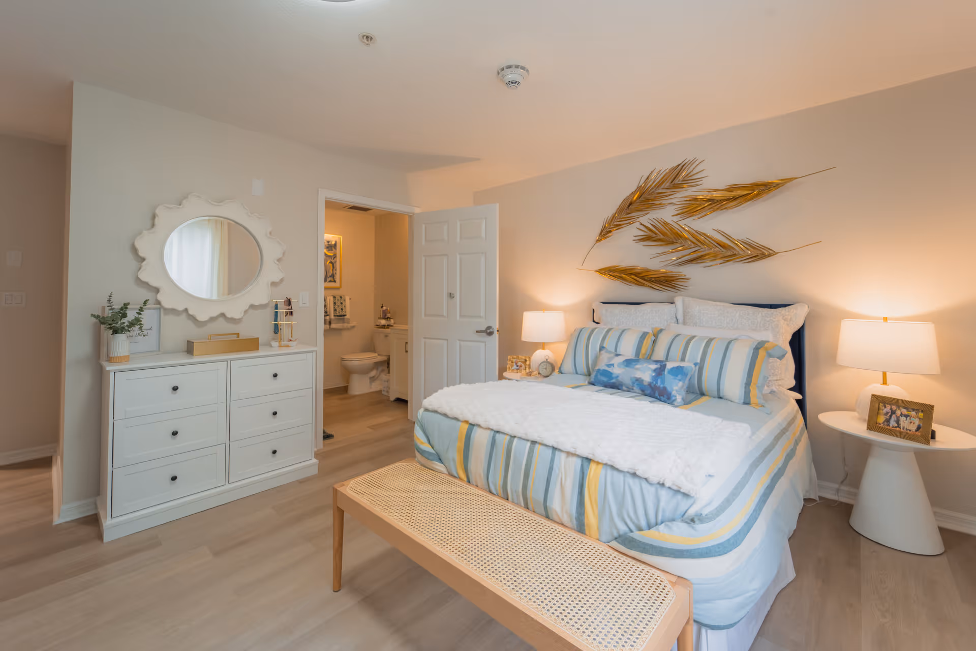 A cozy bedroom with a neatly made bed featuring striped bedding in blue, yellow, and white. There are two bedside tables with lamps and a framed photo on the right table. Above the bed, there is a decorative wall art with golden palm leaves. To the left, there is a white dresser with a round decorative mirror above it and some small decor items. An open door reveals a bathroom with a toilet and a sink.