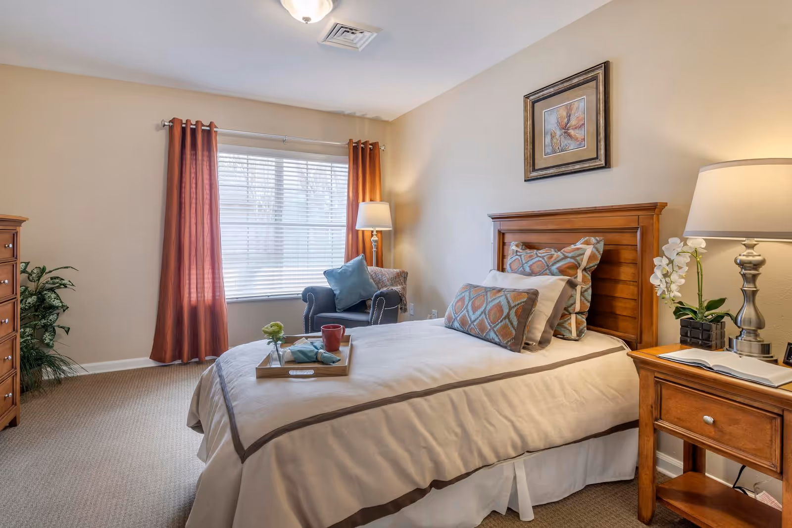 A cozy furnished bedroom with a single bed, wooden headboard, bedside table and lamp, an armchair by a window with curtains, and decorative accents.