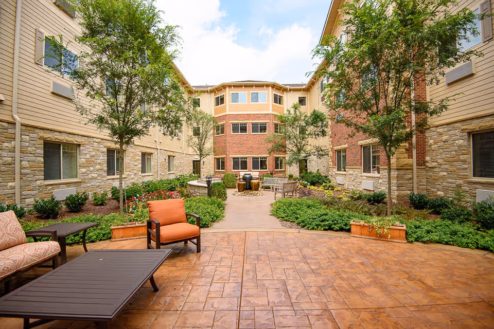 Outdoor courtyard area at The Bridge at Hickory Woods featuring a paved patio with cushioned chairs and tables, surrounded by landscaped greenery, small trees, and a three-story building with brick and stone exterior walls.