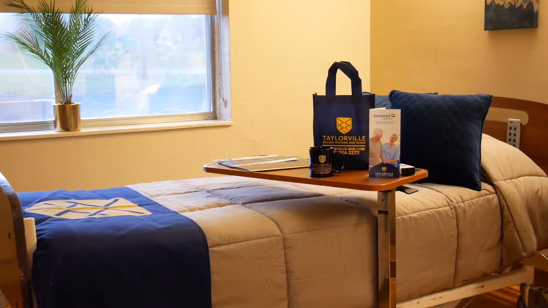 A neatly made hospital-style bed with beige and blue bedding in a room with a window and a potted plant on the windowsill. On the overbed table, there is a blue tote bag, a coffee mug, and a brochure, all branded with Taylorville Skilled Nursing and Rehab logos.