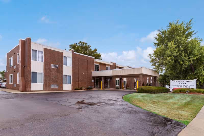 Exterior view of a two-story brick and beige panel building with a covered entrance and a driveway. There is a green lawn with a tree and a sign that reads 'Cambridge East Health Care Center' with a phone number. The sky is partly cloudy.